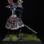 Bunraku de combat Une figurine d’Okko Chronicles, Bunraku de combat, très grande marionnette mécanique vue de derrière, équipée d’une armure et d’une grande masse.