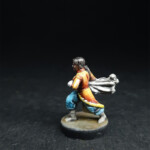 Une figurine du 7ème, La Traqueuse, vue de côté, habillée de vêtement coloré jaune et orange, porte un filet dans ses mains.