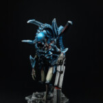 Intrus Adulte V1 Une figurine de Nemesis, Un Intrus adulte version 1, créature vue de face, bipède, possède deux bras et une queue, de couleur bleue pour les carapaces, se tient debout et prend appui sur un débris de fer.