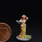 Une figurine du 7ème Continent à côté d’une pièce d’un centime d’euro, la Botaniste, vue de face, une femme en robe jaune qui tient son chapeau d’une main et un livre de l’autre.
