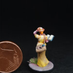 Une figurine du 7ème Continent à côté d’une pièce d’un centime d’euro, la Botaniste, vue de derrière, une femme en robe jaune qui tient son chapeau d’une main et un livre de l’autre.