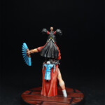 Isako Une figurine de Okko Chronicles, Isako, jeune femme marionnettiste de derrière, vêtue d’une robe rouge, un éventail dans la main et une marionnette dans l’autre.