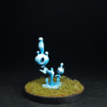 Kami d'eau Une figurine de Okko Chronicles, Kami d’eau, deux petites créatures faites d’eau, une grande et une petite, aux formes arrondies, se tiennent debout dans de l’herbe.