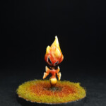 Kami de feu Une figurine de Okko Chronicles, Kami de feu, petite créature faite flamme dans de l’herbe brûlée.